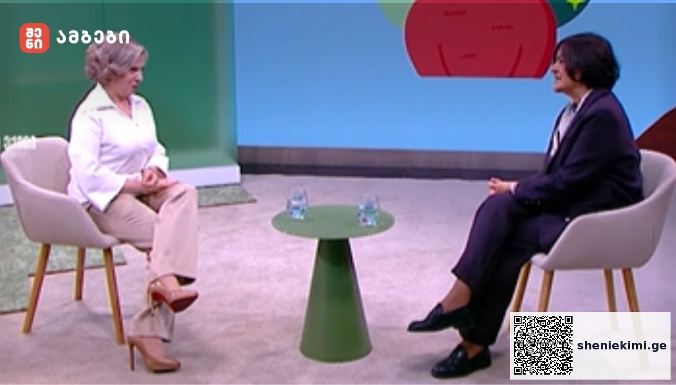 Two people sit in armchairs on a TV talk show set, with a small green table between them and two water glasses on it.