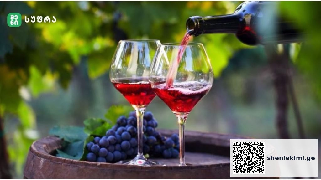 Two red wine glasses on a wooden barrel with wine being poured from a bottle, grapes and vines in the background.