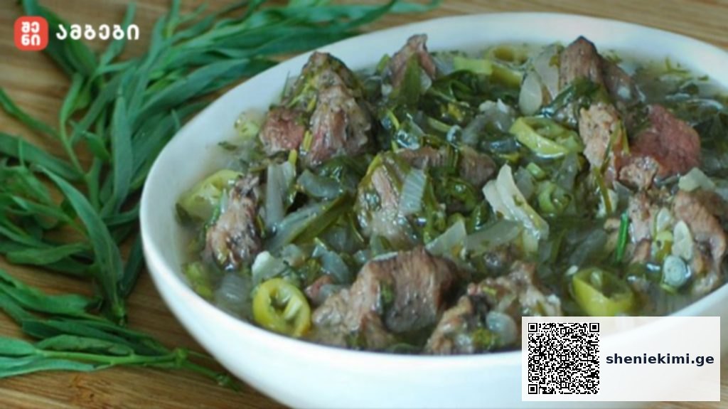 Bowl of beef stew with onions, green peppers and herbs in a light broth on a wooden table, with fresh greens beside it.