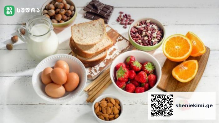 Assorted breakfast ingredients on a white wooden table: eggs, milk, sliced bread, almonds, chocolate, pistachios, strawberries, oats, and orange halves.