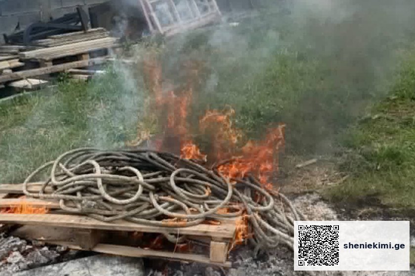 Coiled hoses on a wooden pallet burning in a small outdoor fire with smoke rising nearby debris and grass area on a construction site.