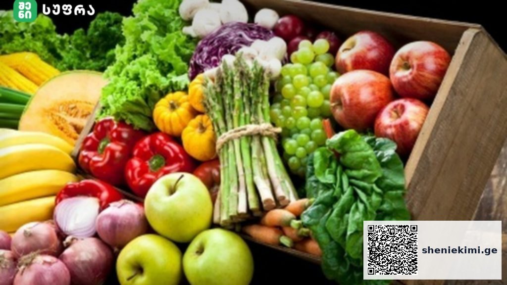 Assorted fresh fruits and vegetables arranged in a wooden crate, including bananas, apples, peppers, leafy greens, grapes, and bunches of asparagus.