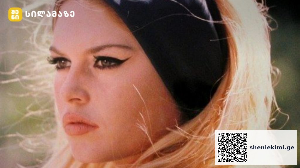 Close-up of a blonde woman with winged eyeliner, wearing a black headband, outdoors in sunlight.