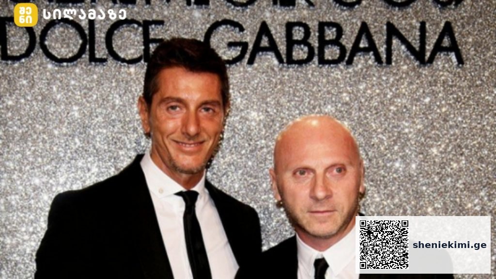 Two men in black tuxedos pose for a photo at a Dolce & Gabbana event, standing before a glittery backdrop.