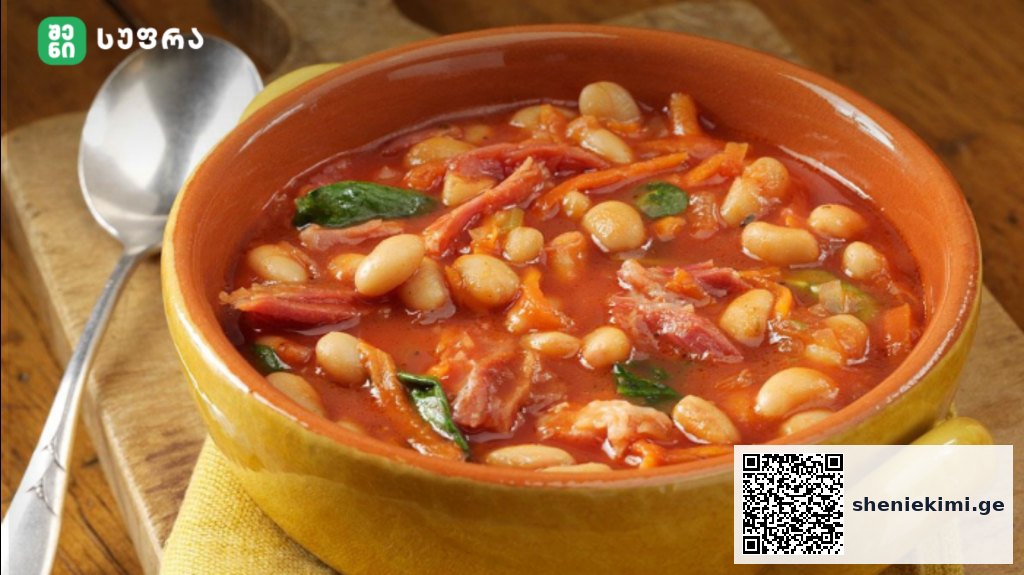 Yellow ceramic bowl holds a hearty white bean and tomato soup with greens on a wooden table, spoon nearby.