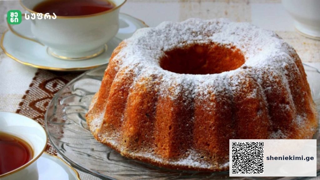 Bundt cake dusted with powdered sugar on a glass platter beside tea cups.