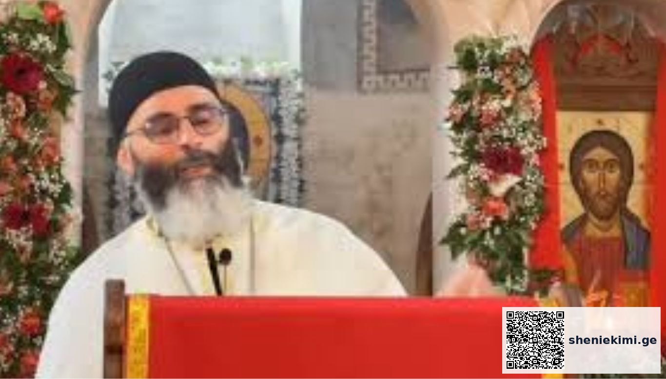 Orthodox priest with a black hat and glasses speaks at a red lectern, with Christ icon and floral garlands nearby.