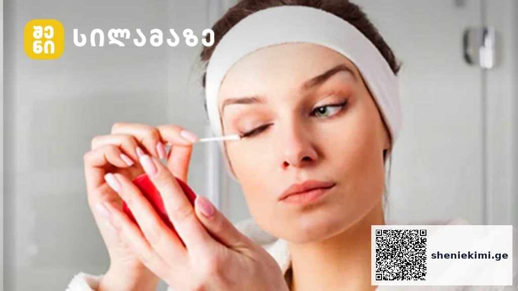 Woman with a white headband applies makeup to her eye using a cotton swab, in a bathroom setting with a logo in the corner.