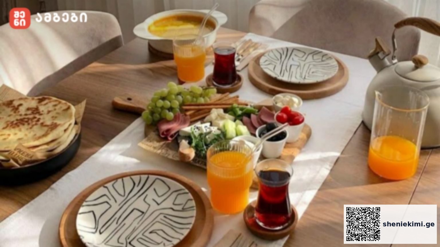 A wooden dining table set for breakfast with a cheese, grape, and meat platter, orange and red drinks, a teapot, and stacked plates nearby.
