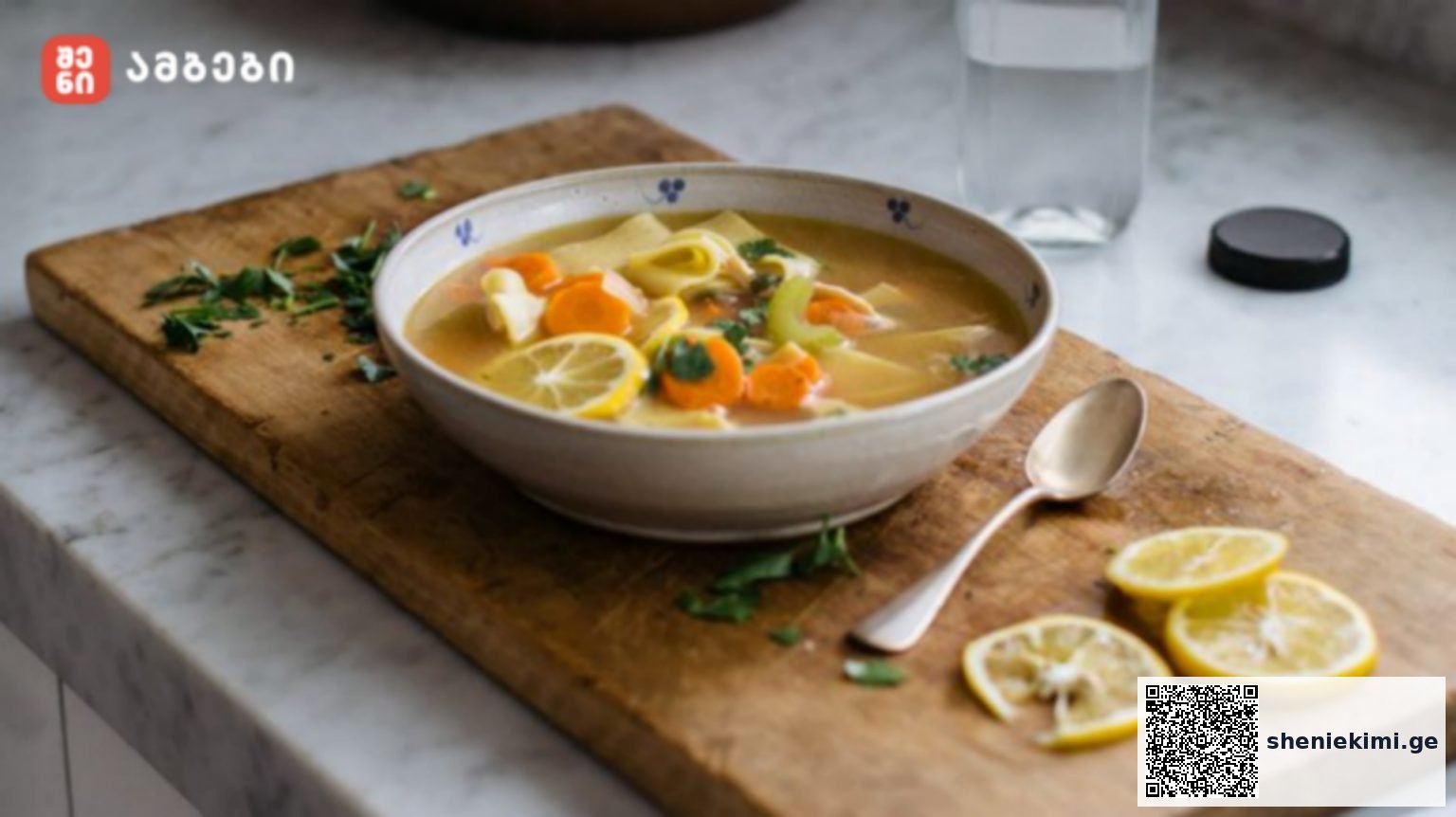 Bowl of lemon chicken soup with carrots and pasta on a wooden cutting board, with lemon wedges and a spoon nearby.