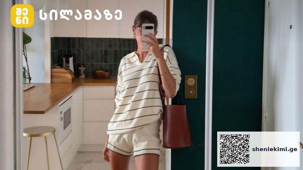 Person taking a mirror selfie in a modern white kitchen, wearing cream striped loungewear and carrying a red bag.
