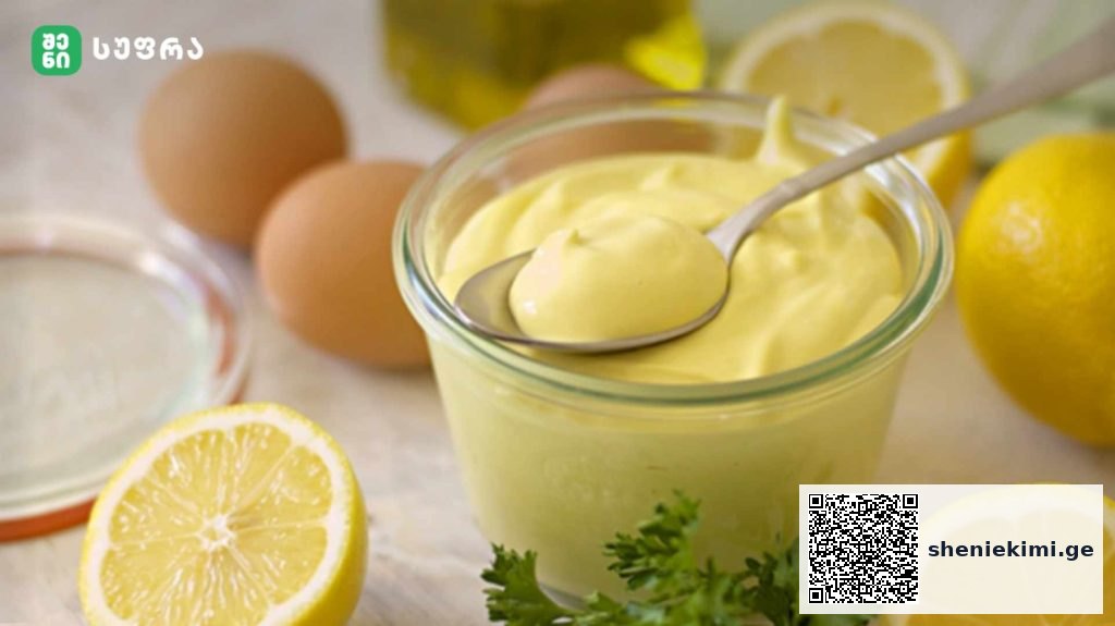Glass jar of creamy lemon mayonnaise with a spoon, surrounded by lemons, eggs, and parsley on a kitchen counter.