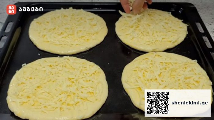 Four round dough discs topped with shredded cheese on a baking sheet; a hand is sprinkling cheese on the top left piece.
