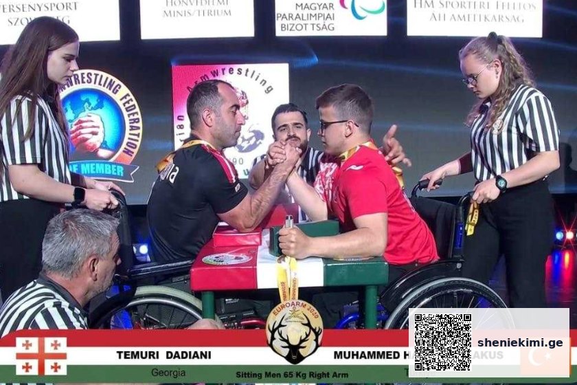 Two wheelchair arm wrestlers face off on a stage, gripping hands as referees observe; banners and flags of Georgia and Turkey appear in the background.