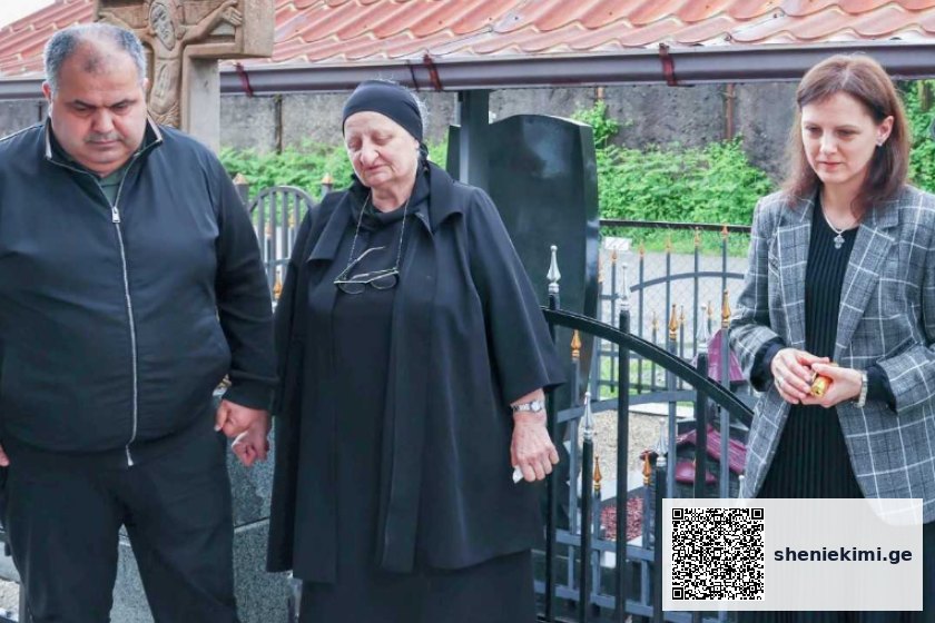 Three adults stand by a grave in a cemetery; a man and an elderly woman in black hold hands, while a woman in a checkered blazer stands nearby holding her hands together.
