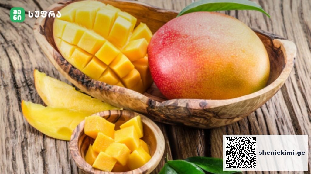 Mango cut into cubes in a wooden bowl beside a whole mango on a rustic wooden surface with green leaves nearby