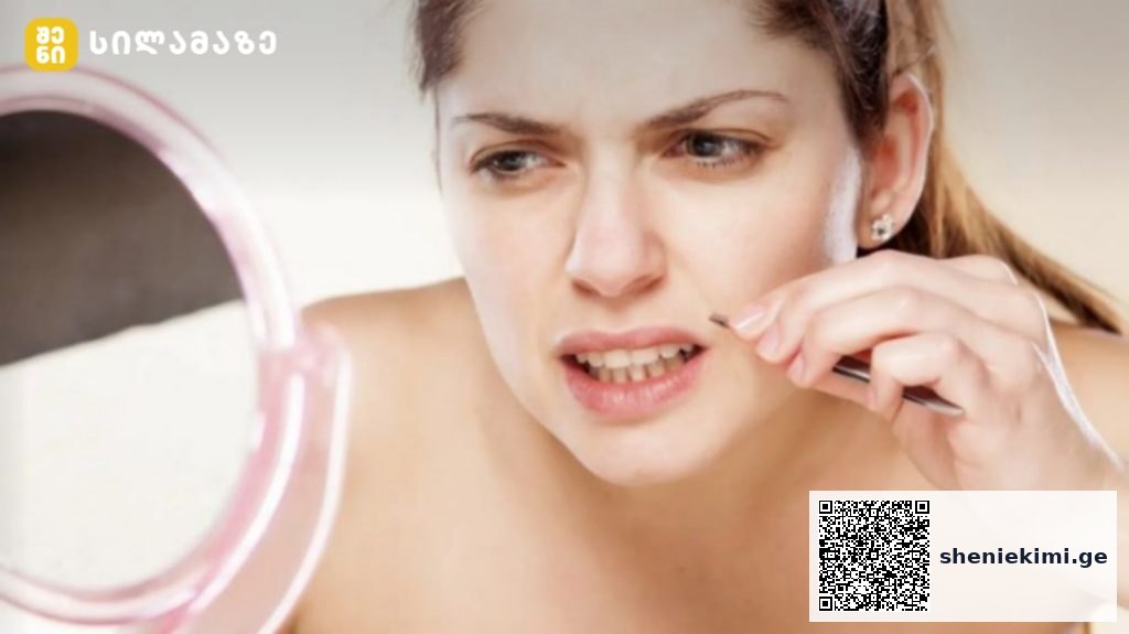Woman plucking eyebrow hair with tweezers while looking in a round pink mirror to her left.