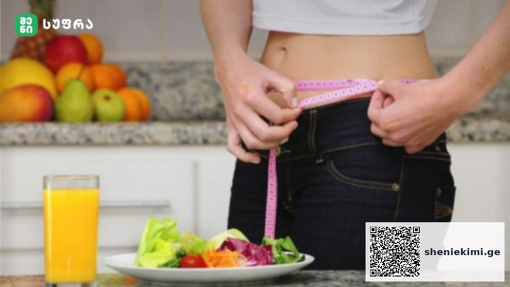 Person in a kitchen measures their waist with a pink tape measure beside a salad plate and glass of orange juice.