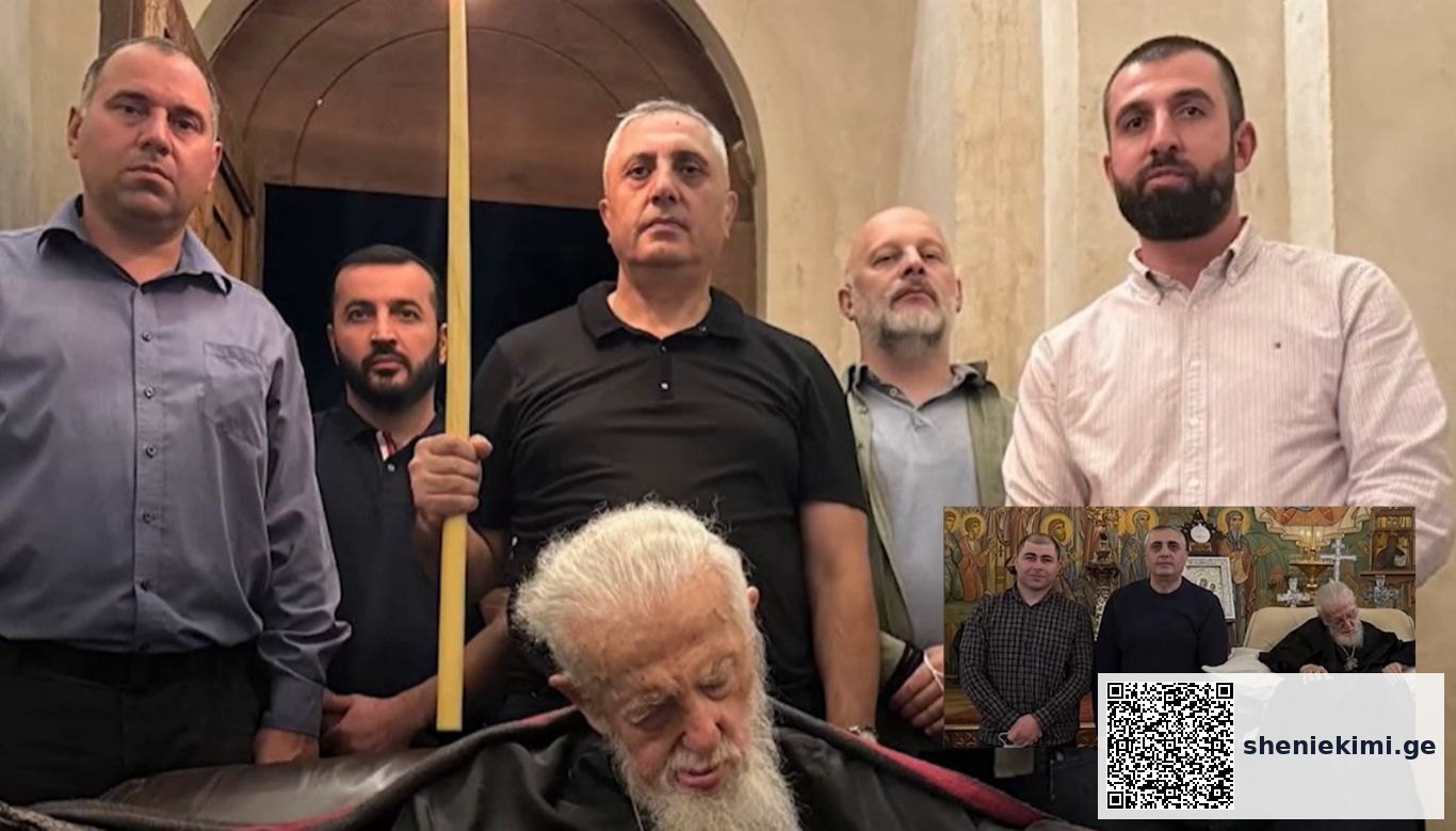 Five men stand behind an elderly man with a white beard lying in a coffin inside a church setting; one man holds a long yellow staff.