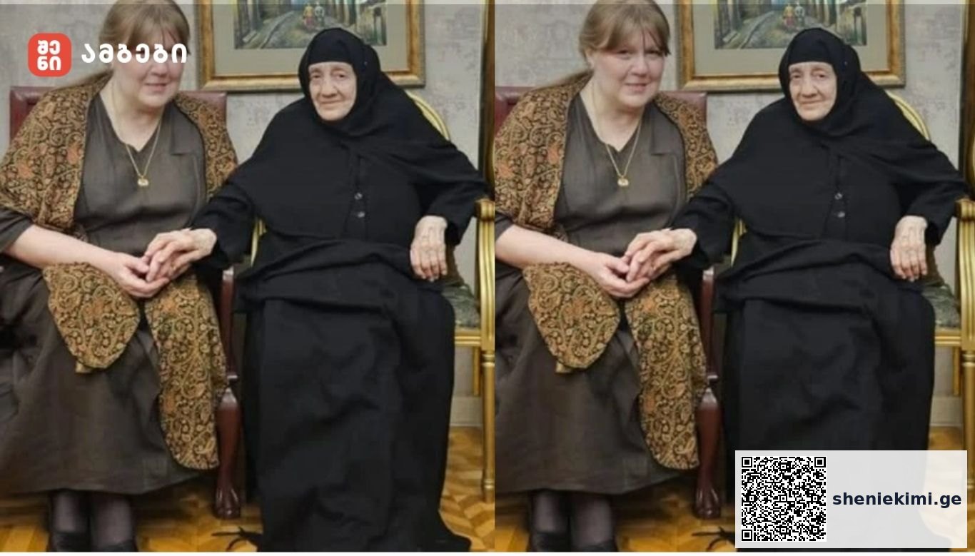 Two women sit indoors on chairs, one wearing a black hijab and dress and the other in a brown patterned shawl, holding hands.