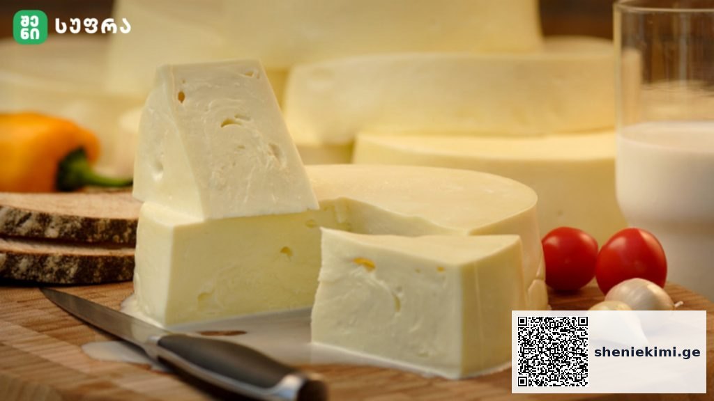 Block of soft cheese with wedges on a wooden cutting board, knife, tomatoes, garlic, and milk in the background.
