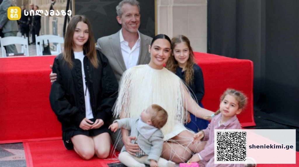 A woman in a white fringe dress sits on a red carpet surrounded by children and adults, posing for a photo at an event.