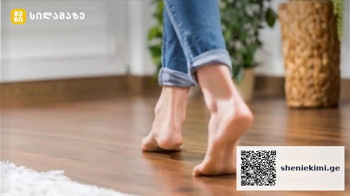 Barefoot person walking on a wooden floor indoors, jeans cuffs rolled up.