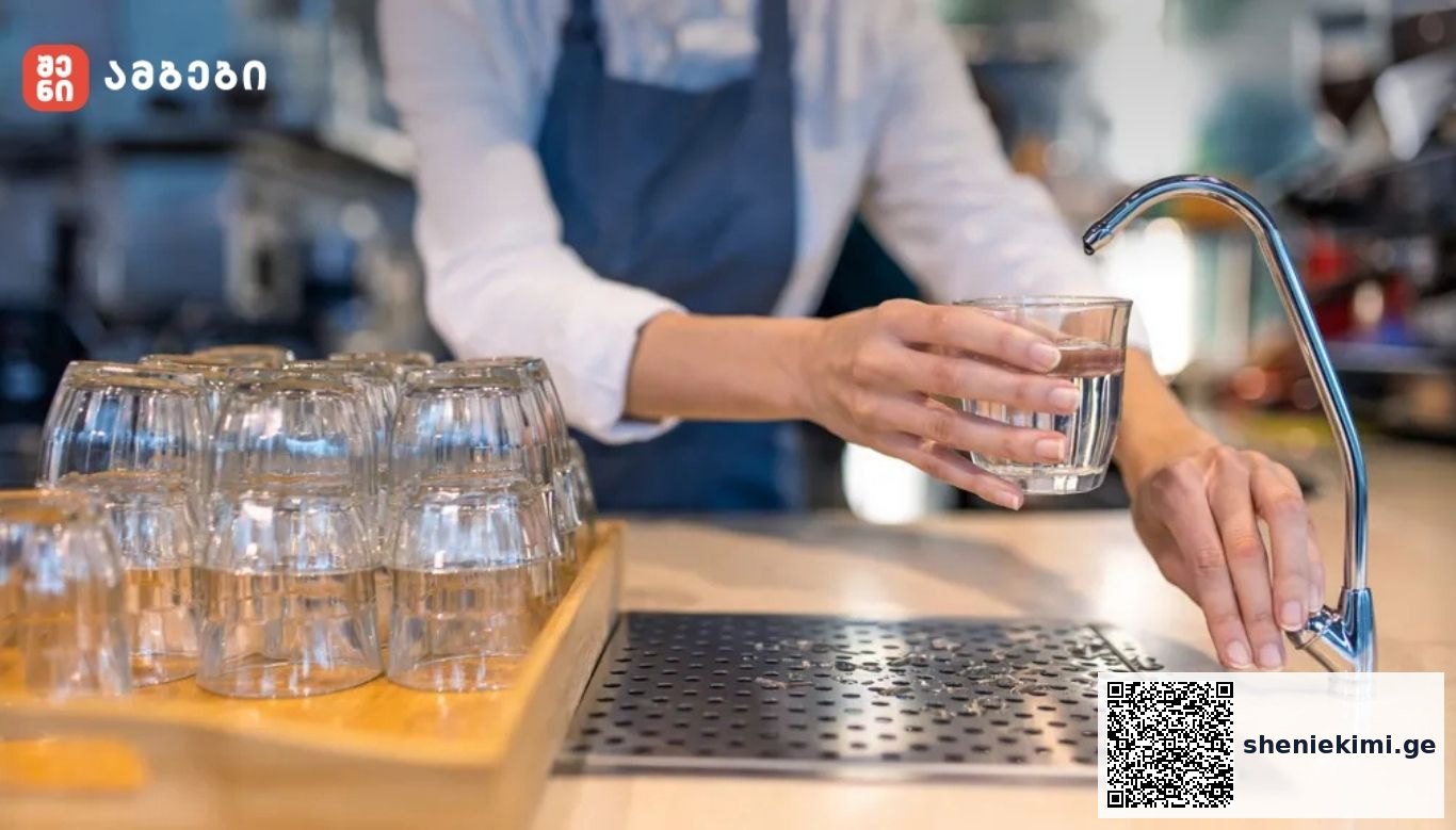 Person rinsing a glass under a kitchen faucet with a rack of glasses nearby on a wooden tray