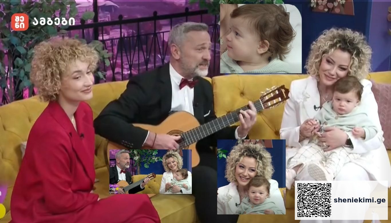 A man in a tuxedo plays an acoustic guitar on a colorful TV talk-show set, flanked by two smiling women holding a baby on a yellow couch.