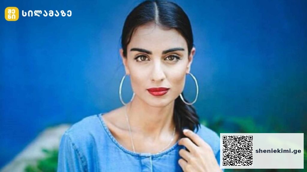 Portrait of a woman with dark hair in a ponytail, wearing large hoop earrings and a blue denim top against a blue background.