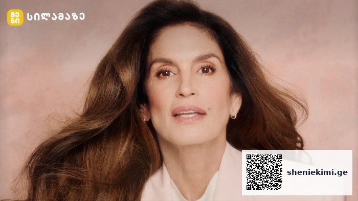 Portrait of a mature woman with long brown hair wearing a white blazer, posed against a peach background with a small logo in the top-left corner.