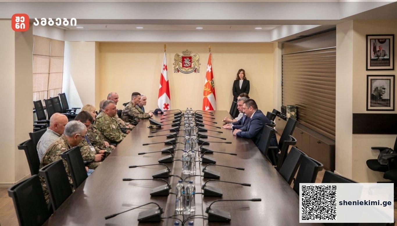Long conference table with military personnel on the left and civilian officials on the right, two flags and a crest behind, plus a woman standing at the far end.