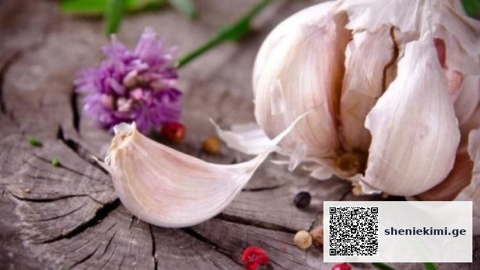 Garlic bulb with a peeled clove on a weathered wooden surface, with a purple flower in the background.