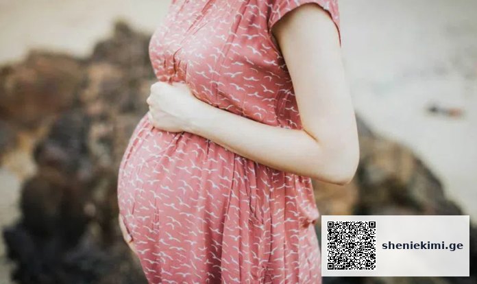 Pregnant woman in a pink patterned dress gently cradling her baby bump outdoors.