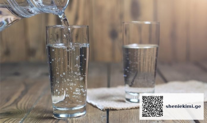 Water is being poured into a tall glass on a wooden table, with a second filled glass nearby and a small towel underneath.