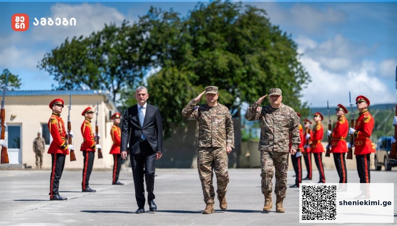 Civilian official in a dark suit walking between two soldiers in camouflage as red-uniform ceremonial guards stand at attention outdoors during a military honor ceremony.