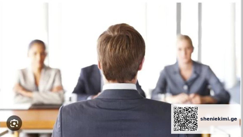 Job interview: a candidate in a suit seated with his back to the camera, facing three interviewers at a table.