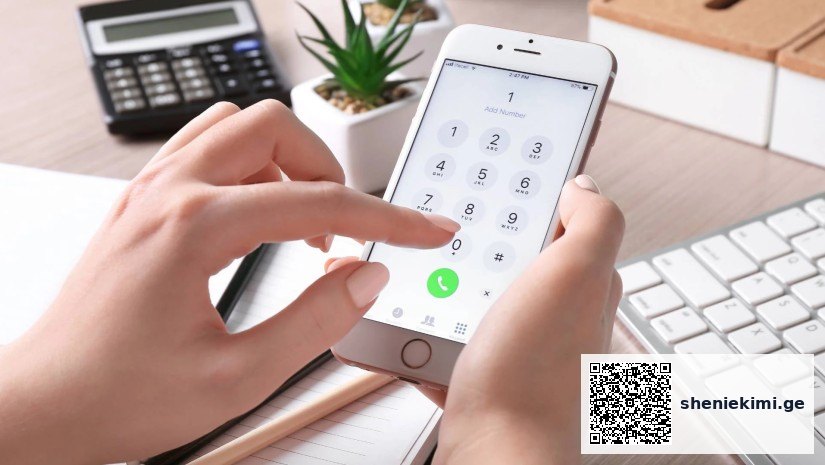 Person holding an iPhone and dialing a number on the phone keypad at a desk with a calculator and plant in the background