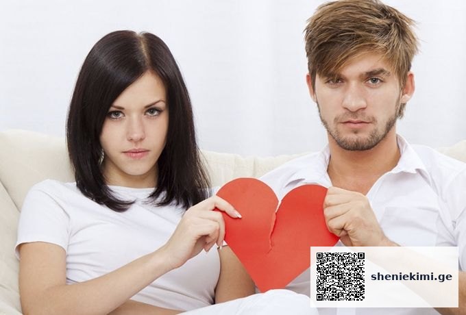Two adults sit on a sofa, holding a torn red paper heart between them, looking unhappy.