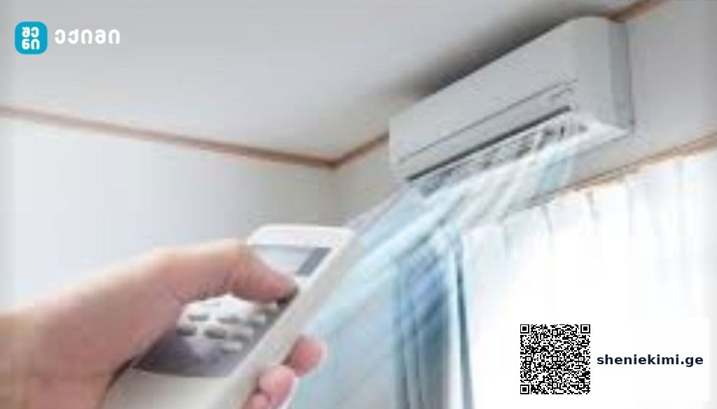 Person holds a remote control aimed at a wall-mounted air conditioner unit above a window with sheer curtains in a home setting.