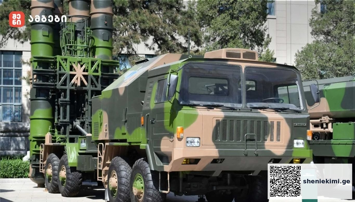 Camouflage military missile launcher on a large six‑wheel truck displayed outdoors in a yard.