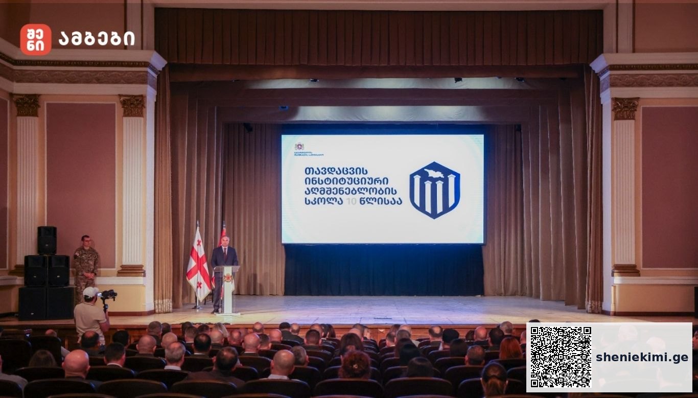 Speaker at a podium on a stage in an auditorium, two Georgian flags beside him and a large screen with a blue emblem and Georgian text behind him