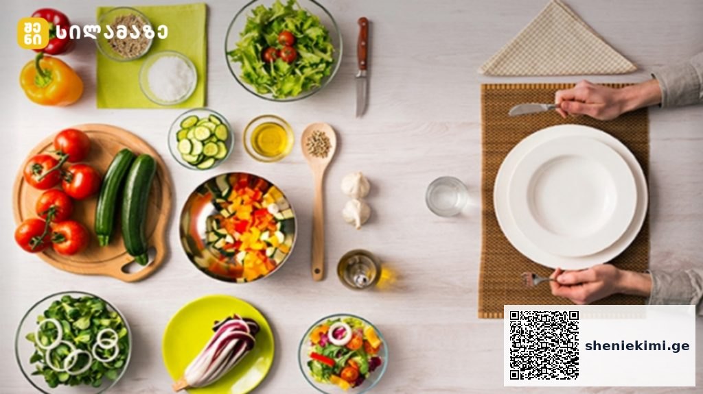 Top-down view of a colorful kitchen spread with tomatoes, cucumbers, peppers, bowls of chopped veggies, and a person setting a plate at the table