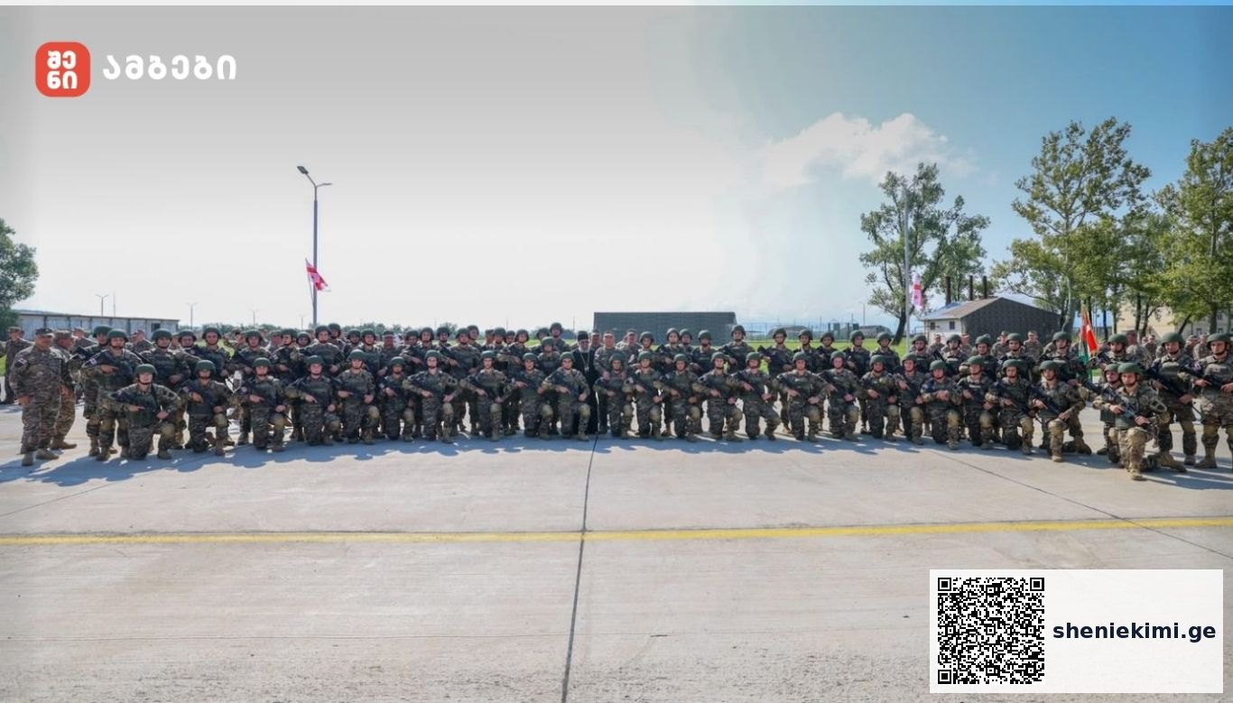 Large group of soldiers in camouflage uniforms posing in a line on a tarmac, with trees and a building in the background.