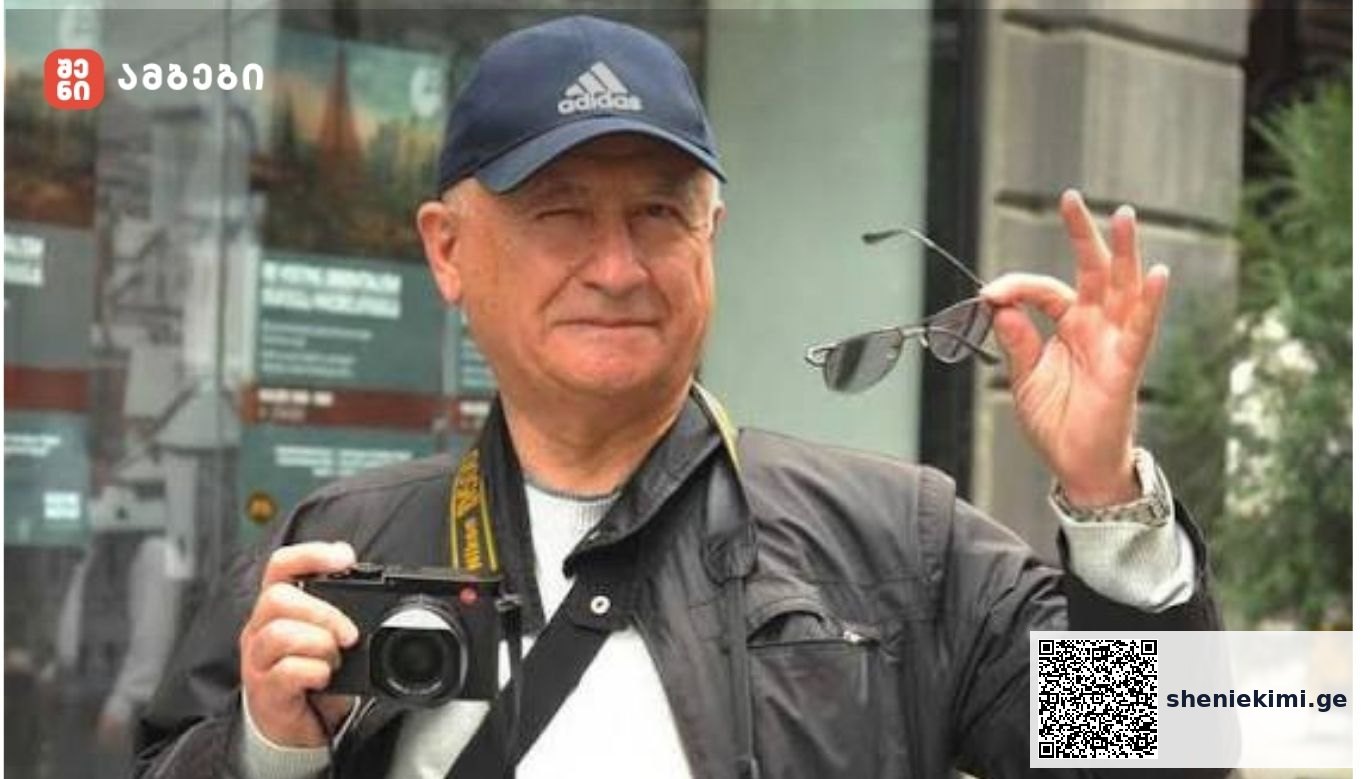 Older man wearing a blue Adidas cap holds a camera in front of him and lifts sunglasses in his right hand.
