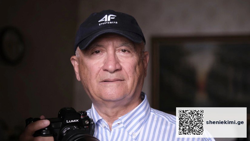 Portrait of an older man in a striped shirt wearing a cap, holding a camera with a Lumix lens.
