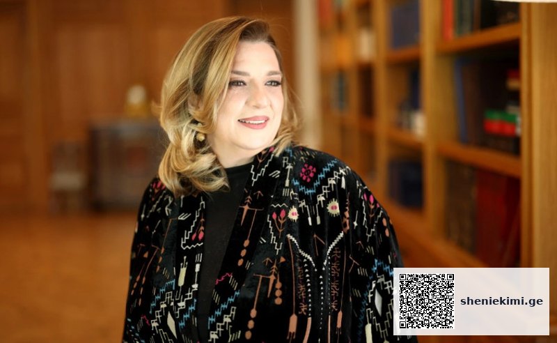 Portrait of a smiling woman with blonde hair wearing a patterned black coat, standing in a library with bookshelves in the background.