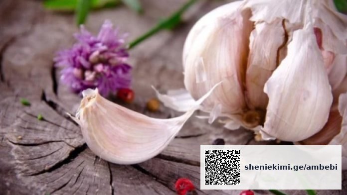Garlic bulb with a peeled clove on a weathered wooden surface, with a purple flower in the background.