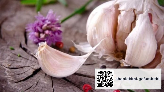 Garlic bulb with a peeled clove on a weathered wooden surface, with a purple flower in the background.