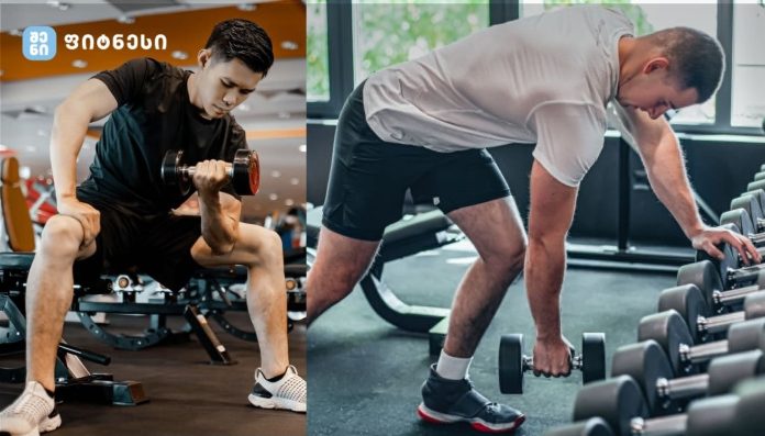 Two men in a gym: one bench-pressing a dumbbell, the other bending to pick up a dumbbell from a rack.